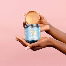 A hair mask in a clear gel like texture in a blue tab featured on a pink background with a woman's hands holding the product.