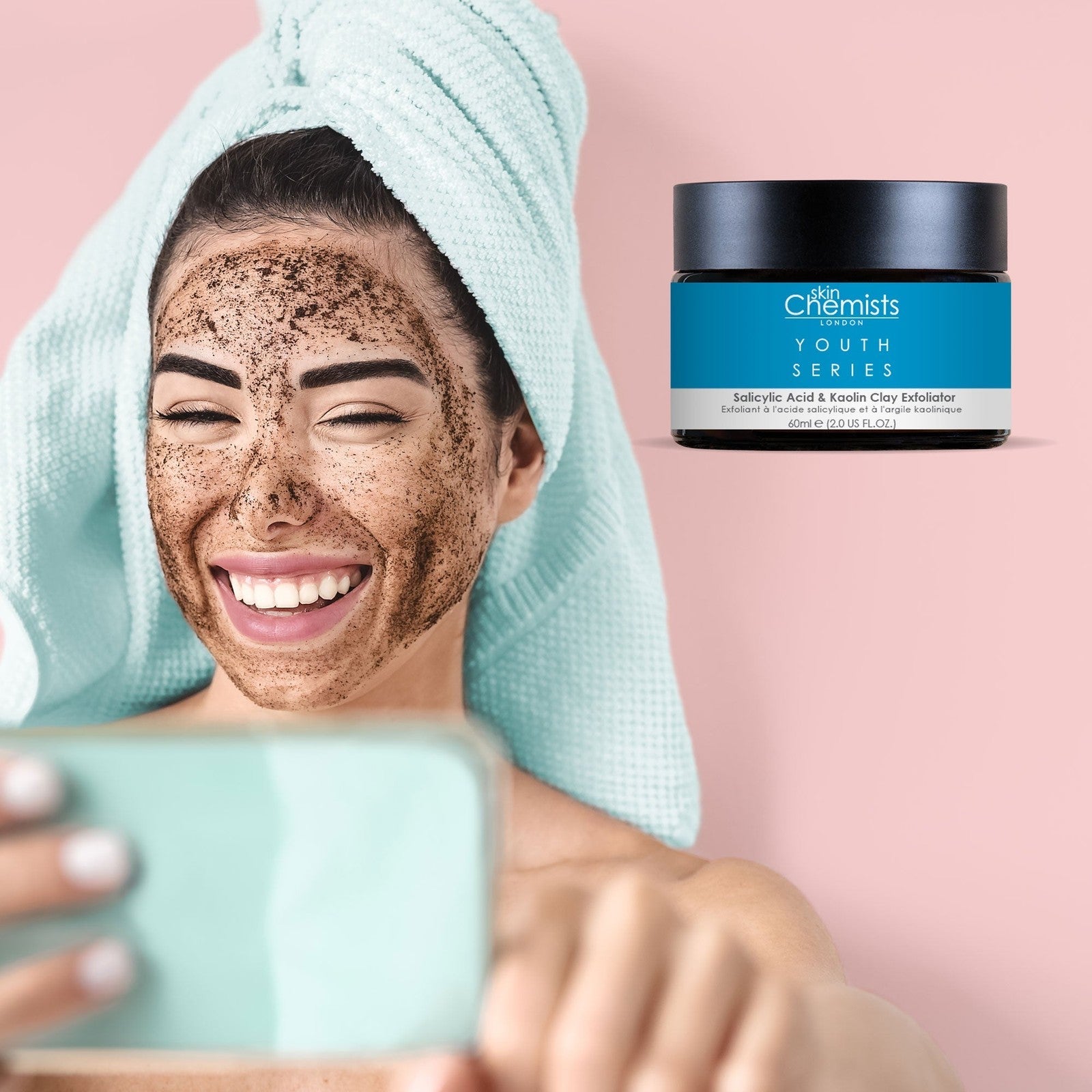 Woman with a scrub on her face using a smartphone, with a jar of skincare product in the corner.