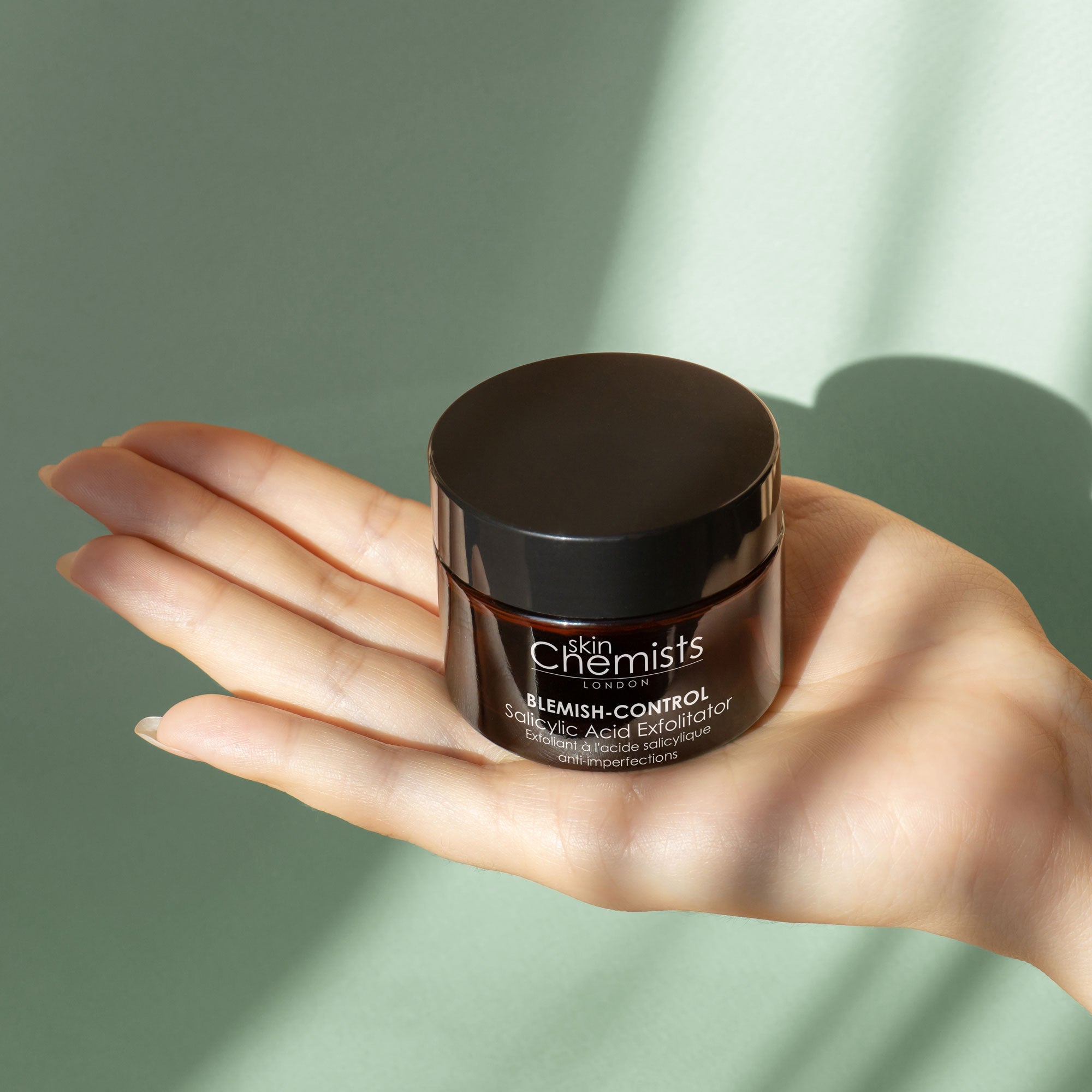 Hand holding a jar of Skin Chemists skincare product against a green background