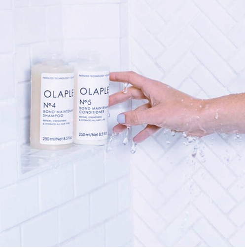 Olaplex shampoo and conditioner bottles held by a hand against a tiled shower wall.