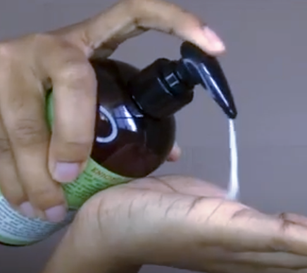 Hand holding a brown spray bottle with a green label, dispensing liquid onto a surface.