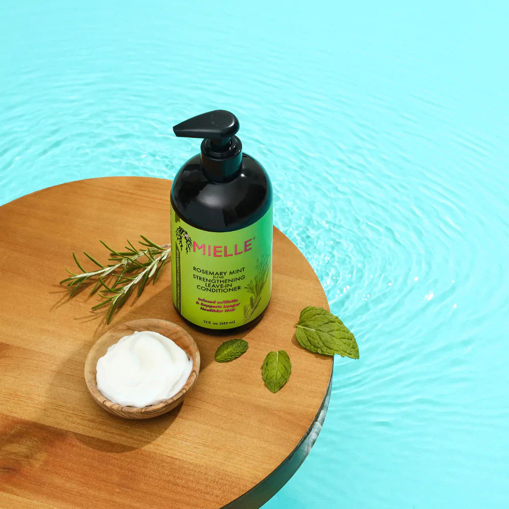 Mielle Organics bottle with green label on a wooden board with leaves and cream, against a blue water background